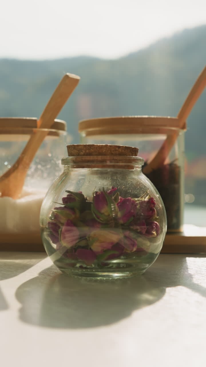 Herbal tea ingredients put on table closeup. Glass jars with dried flowers, herbs and sugar on white countertop by window. Set for organic beverage