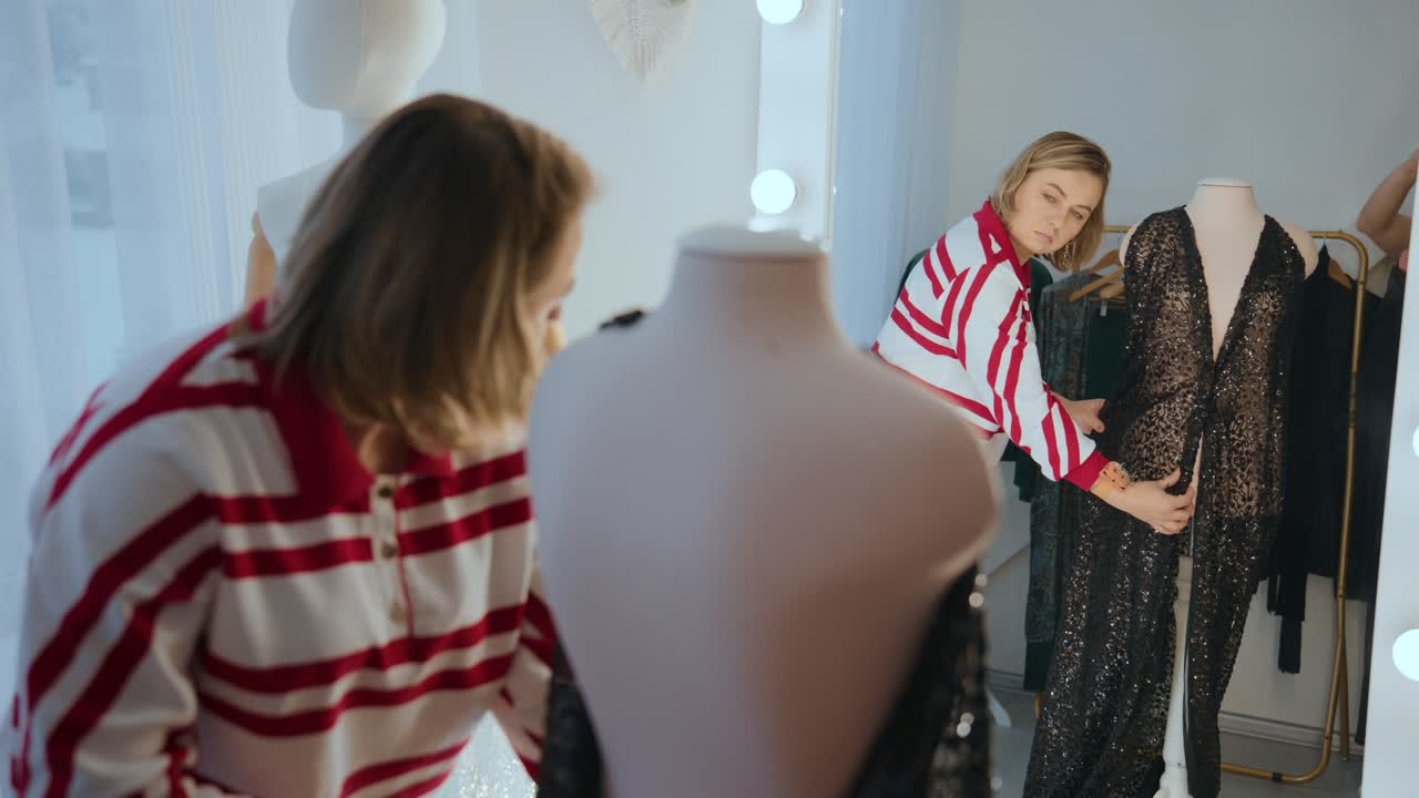 Mulher experimenta vestido de renda preta em um provador iluminado. Ela olha no espelho, verificando a roupa com cuidado e confiança. Manequins estão próximos, vestidos com peças semelhantes