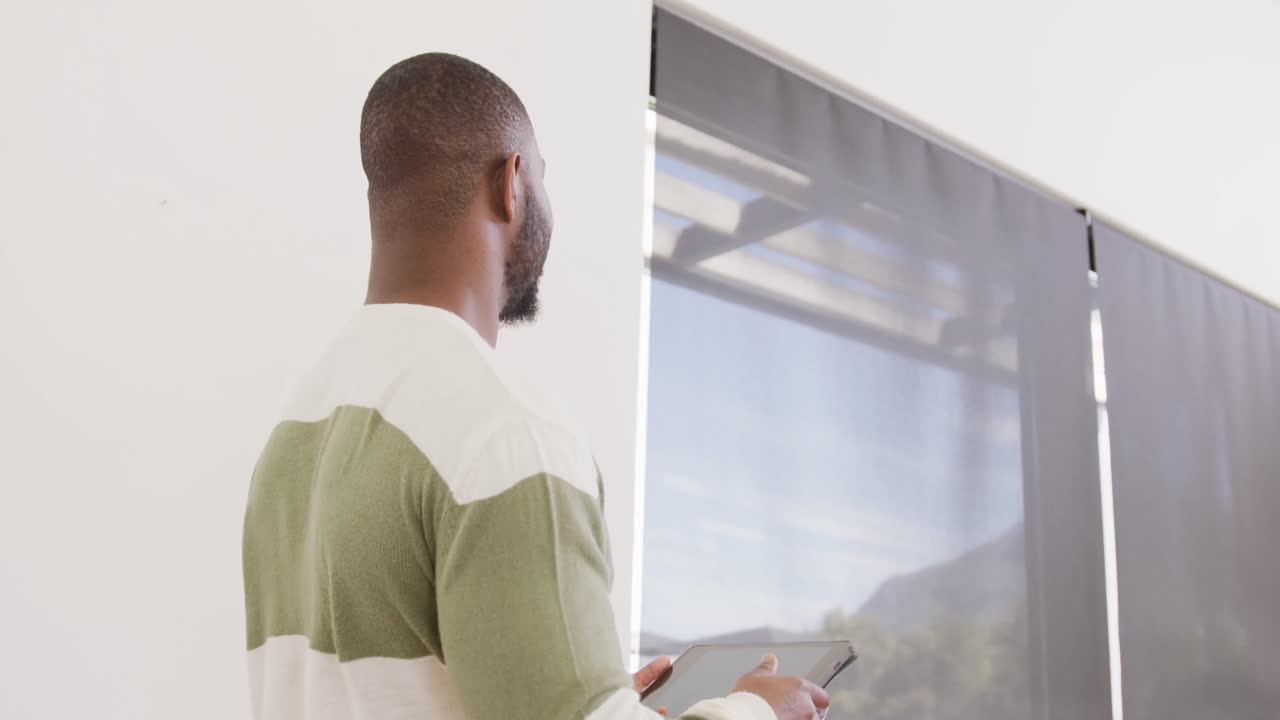 Video of happy african american man smiling, standing using tablet at home