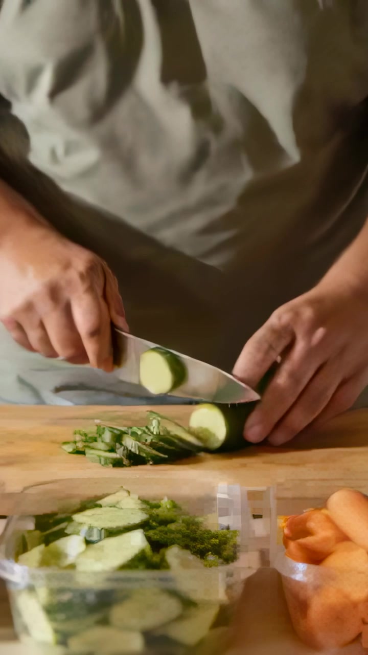 Video verticale: Mani che preparano, con una maglietta verde oliva, affettano un cetriolo sul bancone della cucina con un coltello.