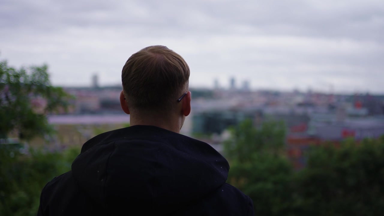 Persona creativa all'aperto, cielo cupo, che si affaccia sulla città, mani che si tolgono il cappuccio, sfondo di natura urbana