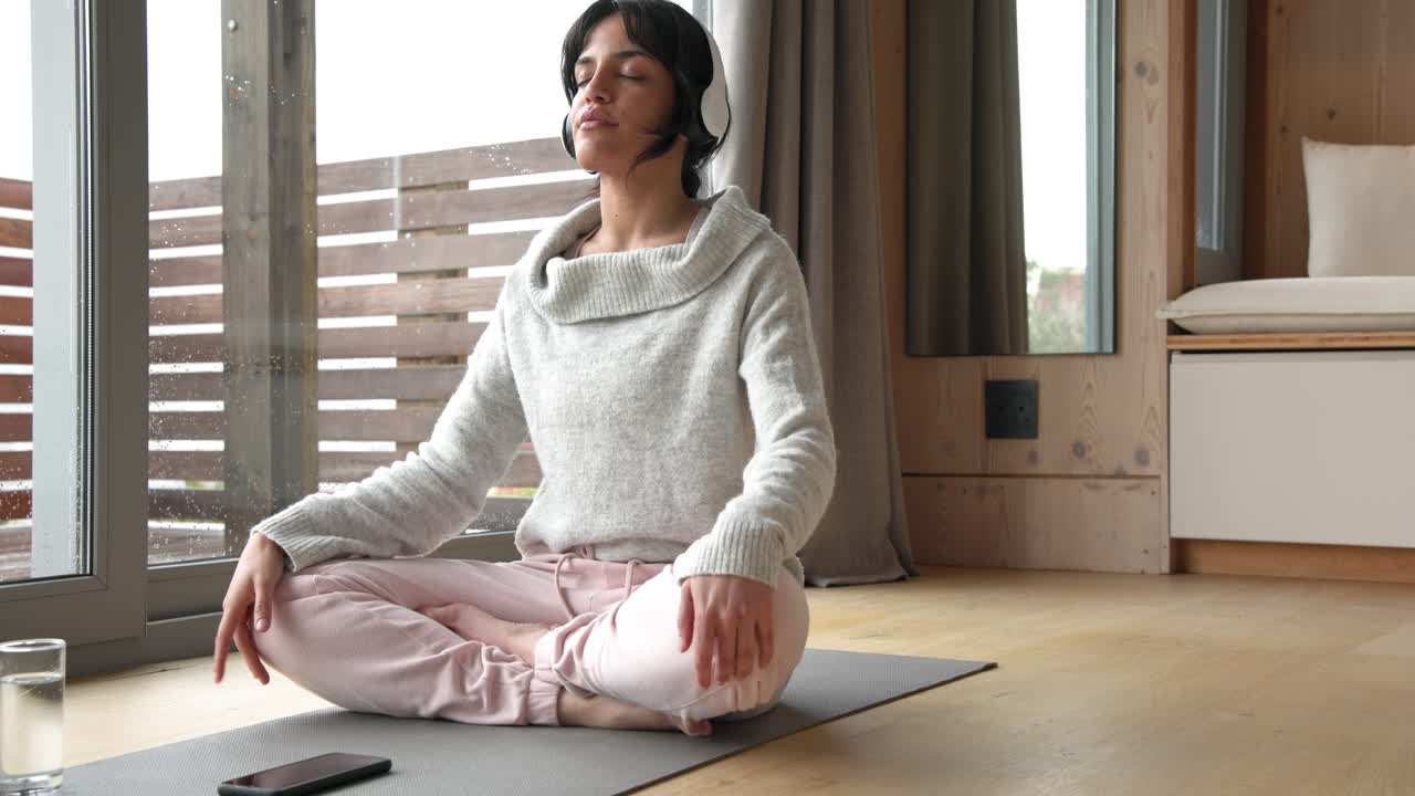 Woman meditating on yoga mat at home, wearing headphones, feeling relaxed