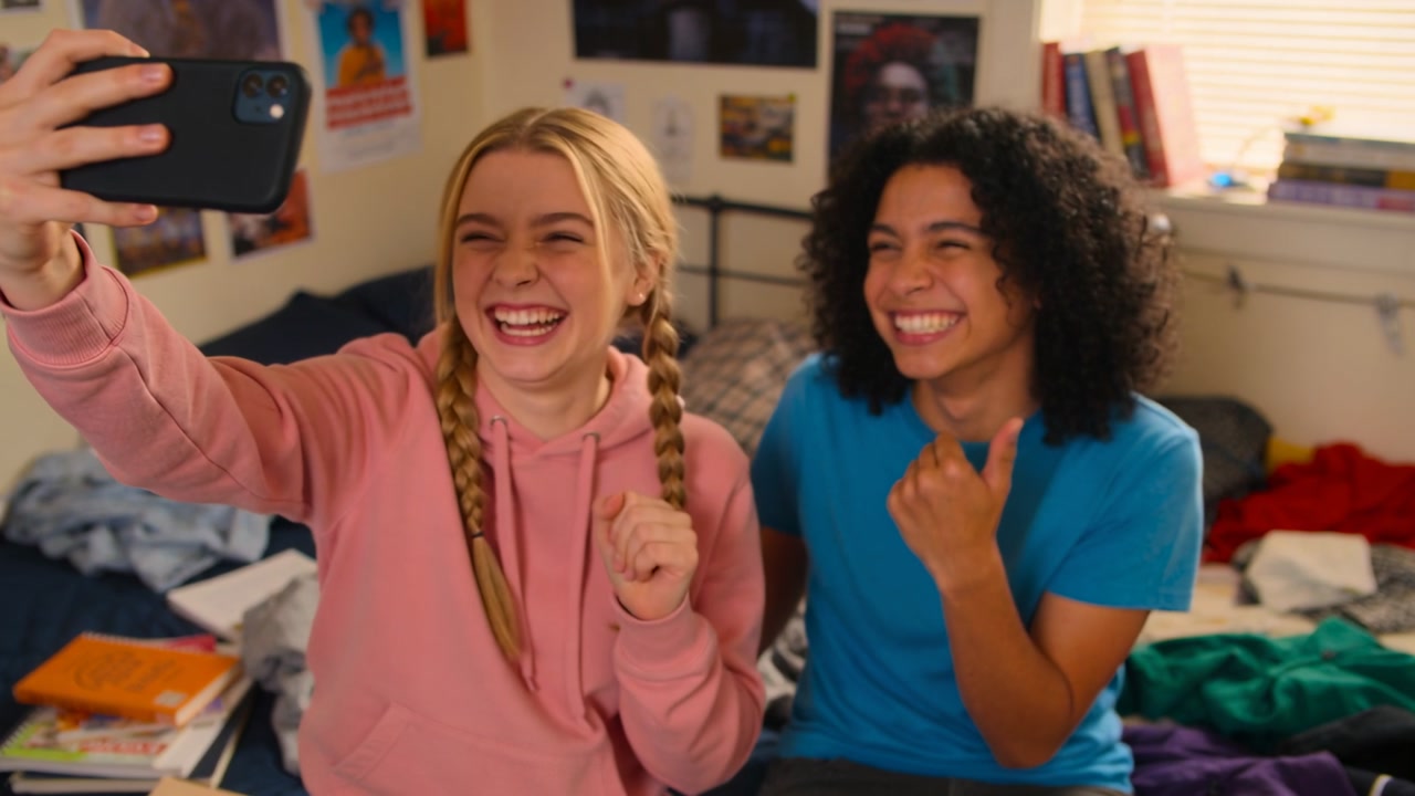 Extending smartphone left teen taking selfies in bedroom, pink hoodie and blue tee laughing for fun