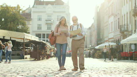 Pareja-Mayor-Caminando-En-El-Centro-De-La-Ciudad-Con-Un-Mapa-Y-Una-Tableta-En-Un-Agradable-Día-Soleado