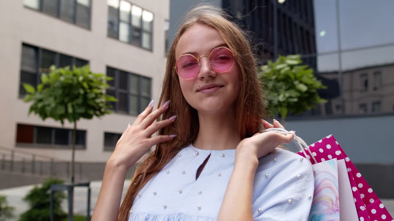joven con gafas de sol rosas de pie frente al centro comercial y sosteniendo bolsas de compras