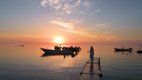 Frau-Im-Weißen-Kleid-Mit-Hut-Läuft-Auf-Dem-Pier-Mit-Wunderschönem,-Farbenfrohem-Sonnenaufgangshimmel-In-Der-Dämmerung-Mit-Spiegelungen-Auf-Dem-Wasser-Und-Schwimmenden-Hölzernen-Fischerbooten-Im-Hintergrund,-Bahrain