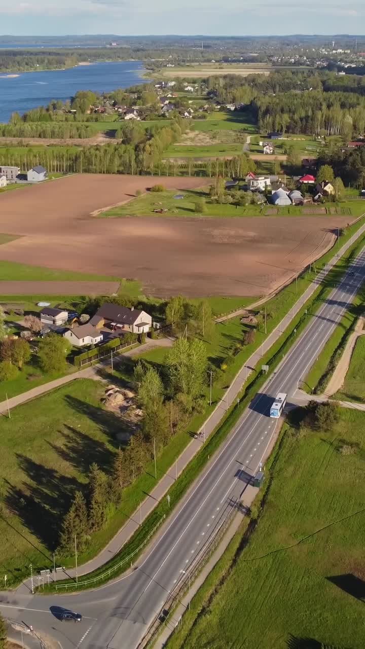 Vertical drone pull away shows Katlakalns spring residential area with houses, Daugmale road traffic, tilled fields, and Daugava river in background under clear sky and afternoon light