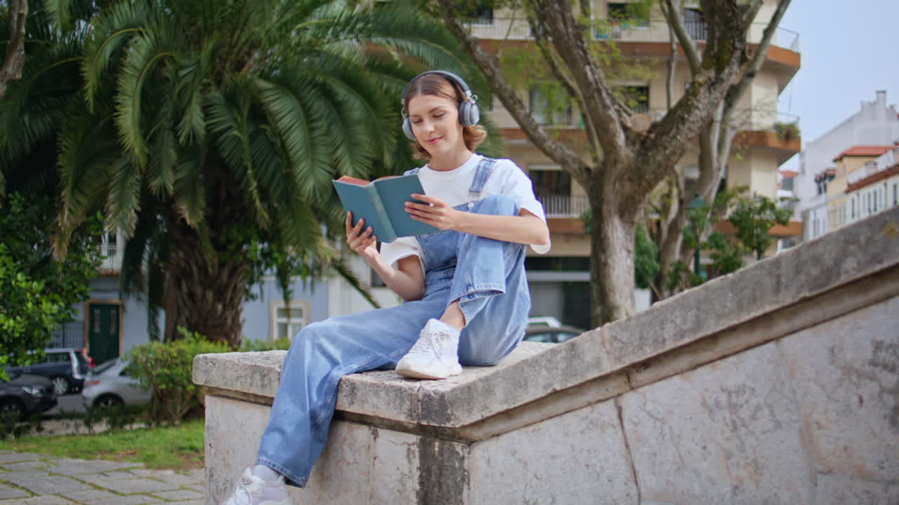 Thoughtful girl reading book in headphones sitting park. Relaxed woman enjoying