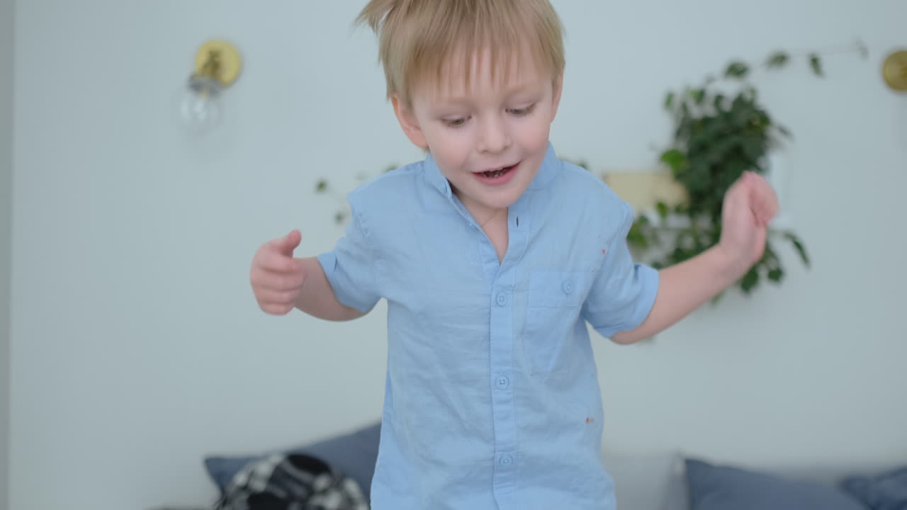 Handsome boy with white hair and blue shirt is jumping on the sofa and looking at the camera in slow motion
