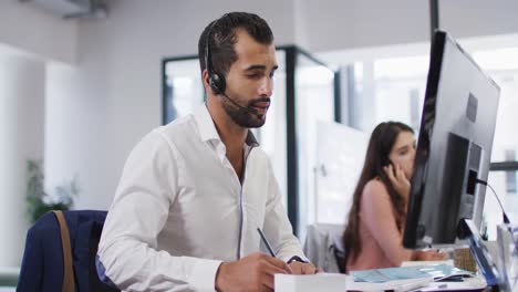 hombre de negocios de raza mixta sentado en un escritorio hablando usando auriculares de teléfono y computadora en una oficina ocupada
