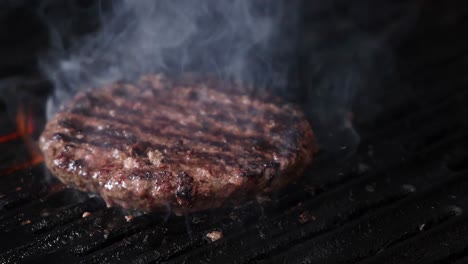 a burger patty sizzles and smokes on a grill, with occasional flare-ups visible.