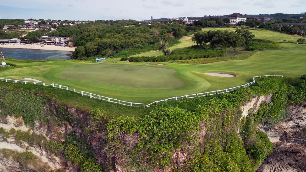 Drone view of a cliff edge golf course with sculpted fairways, clean sand bunkers, cart paths and layered terrain stepping down across a coastal plateau under bright clear daylight in Bali
