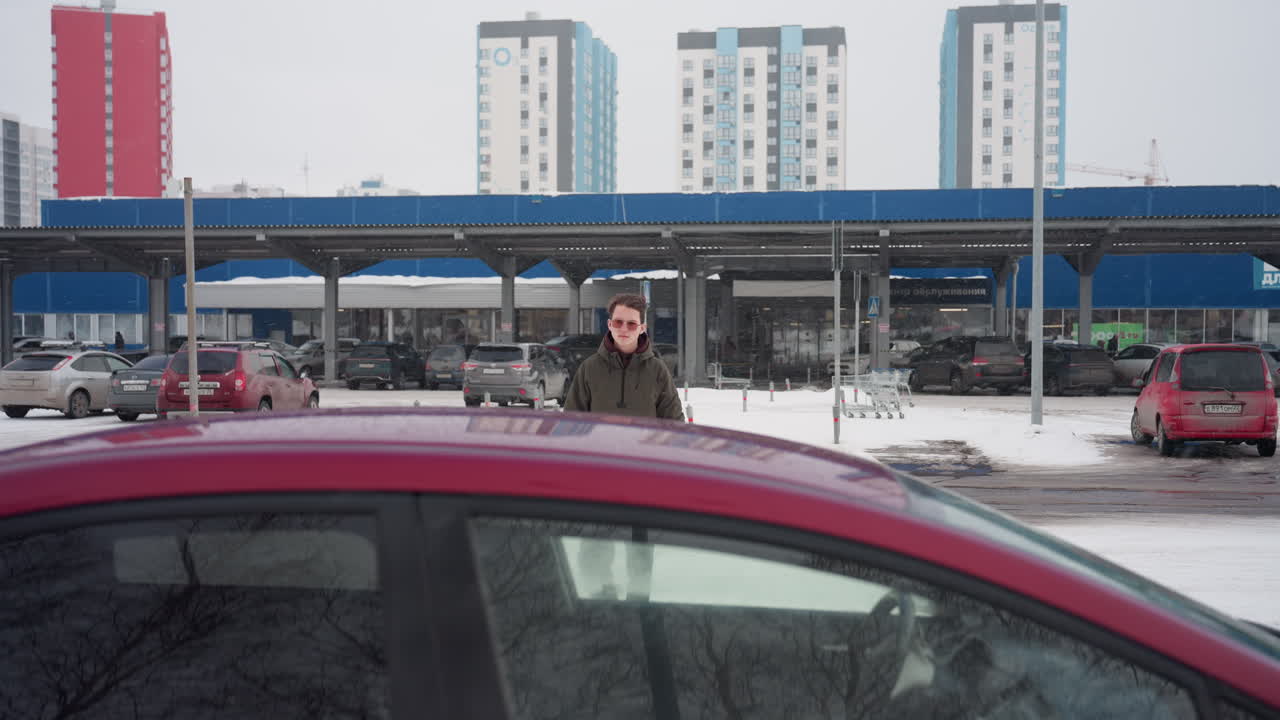 Young man wearing sunglasses walking toward parked red car in snowy parking lot with other parked cars in background and tall residential buildings under cloudy winter sky in urban environment