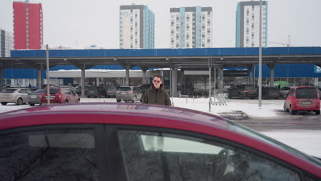 young man wearing sunglasses walking toward parked red car in snowy parking lot with other parked cars in background and tall residential buildings under cloudy winter sky in urban environment