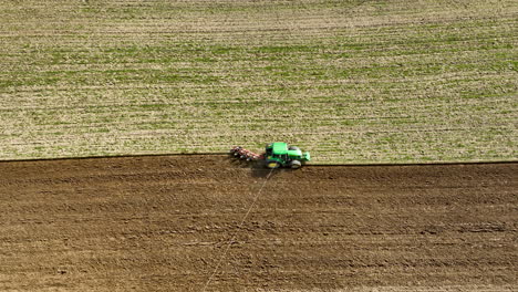 Aerial-close-up-of-a-green-tractor-with-plowing-equipment-working-the-soil-on-farmland,-creating-a-clear-line-between-the-plowed-field-and-the-untouched-green-crop-area