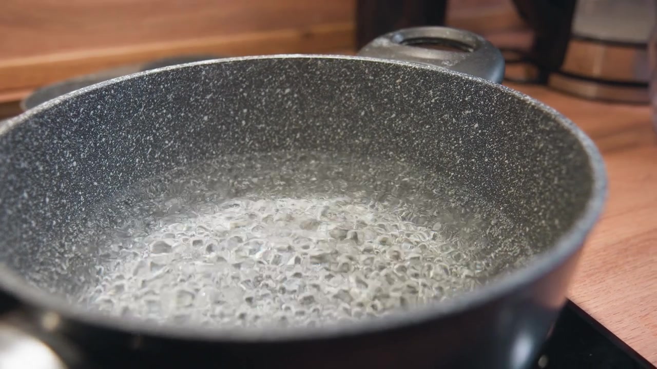 Close-up view of speckled nonstick frying pan on stovetop showing vigorous bubbling and shimmering liquid surface as water or oil reaches active boil, with faint steam and warm kitchen light