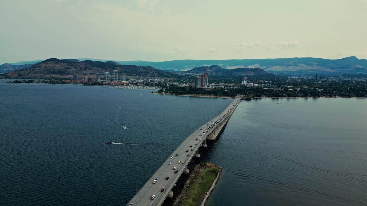 Ripresa aerea al contrario sul William R Bennett Bridge, Okanagan Lake, Columbia Britannica; acqua blu calma, traffico di auto e camion che percorrono il ponte, sfondo montuoso, cielo azzurro chiaro e nuvole rade