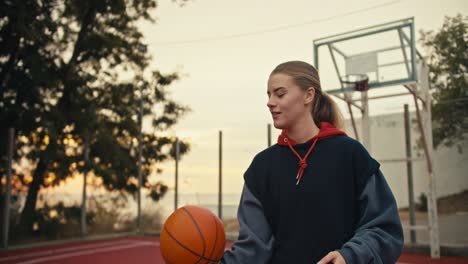 una chica rubia feliz con una sudadera negra trata de hacer girar una pelota de baloncesto naranja en su dedo índice durante su práctica de baloncestro en una cancha de balonceso roja temprano en la mañana