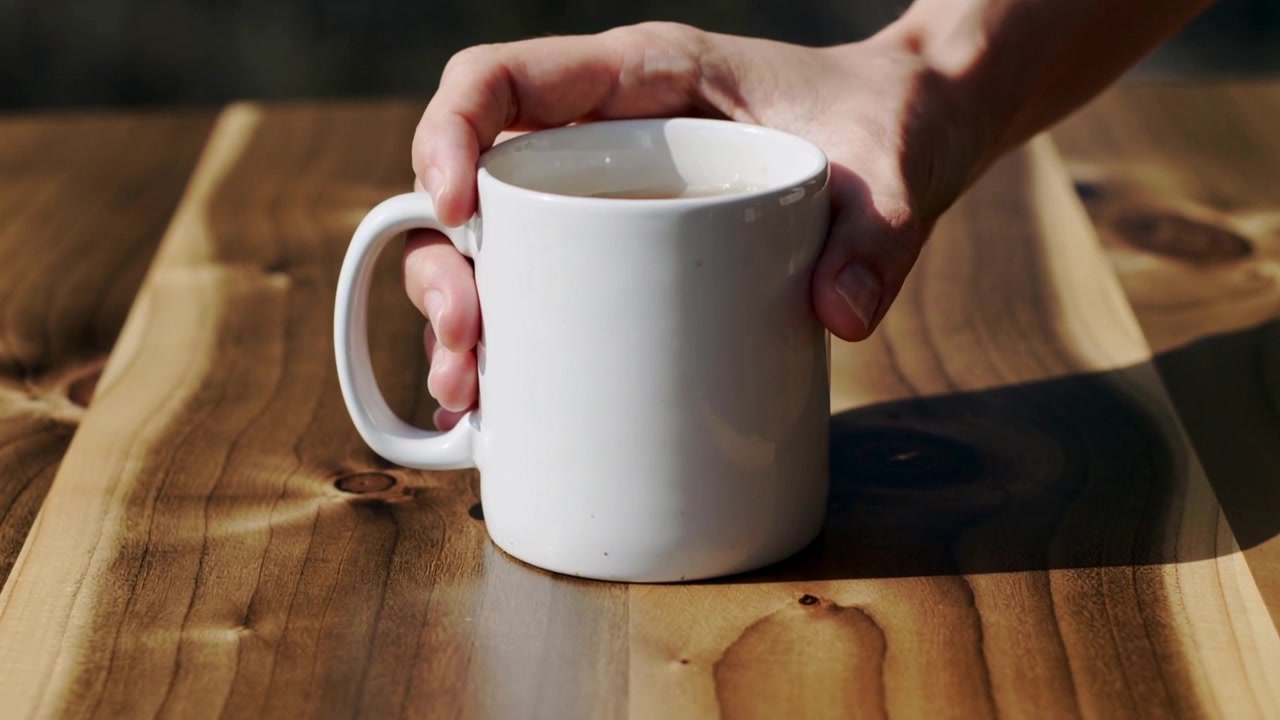 Mãos abaixando e colocando caneca de cerâmica branca em mesa de madeira sob a luz do sol, revelando bebida e sombra.