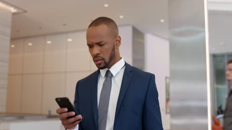 Un-Hombre-De-Negocios-Afroamericano-Revisando-Su-Teléfono-Inteligente-Abrumado-Perdido-En-Un-Lobby-Corporativo-Intimidante