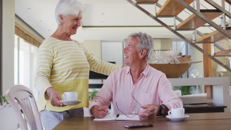 Senior-caucasian-woman-giving-coffee-cup-to-her-husband-while-he-calculates-finances-at-home