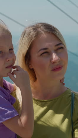 doubting woman with little daughter in arms stands against modern ropeway and old mountains at highland resort on sunny day closeup slow motion