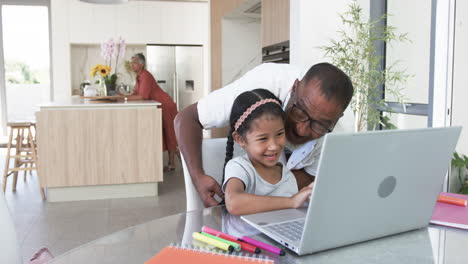 El-Abuelo-Birracial-Ayuda-A-Una-Joven-Nieta-Birracial-Con-Una-Computadora-Portátil-En-Una-Cocina-Moderna