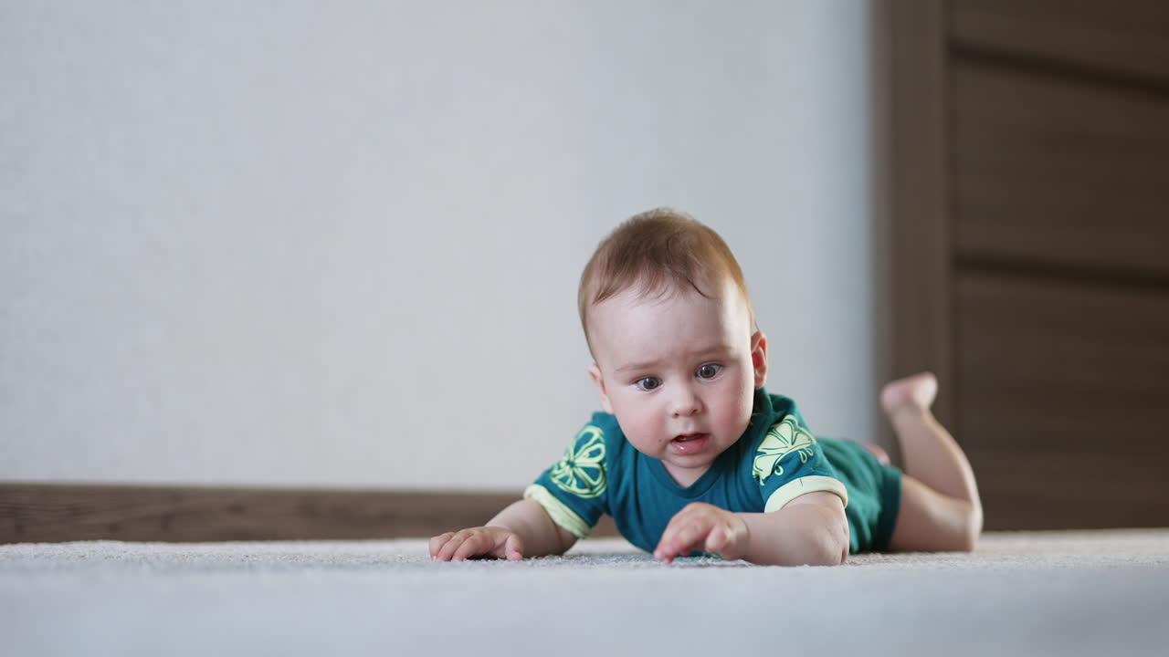 Little Caucasian infant boy lies on the floor banging hands and feet upon it. Cute emotional toddler takes a fist into mouth looking back.