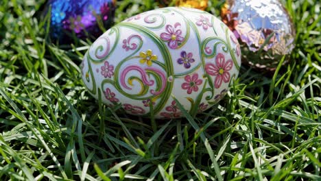 huevo de pascua blanco decorado con patrones florales rosados y verdes, colocado en una cama de hierba verde vibrante, rodeado de huevos de chocolate coloridos envueltos en papel de aluminio brillante