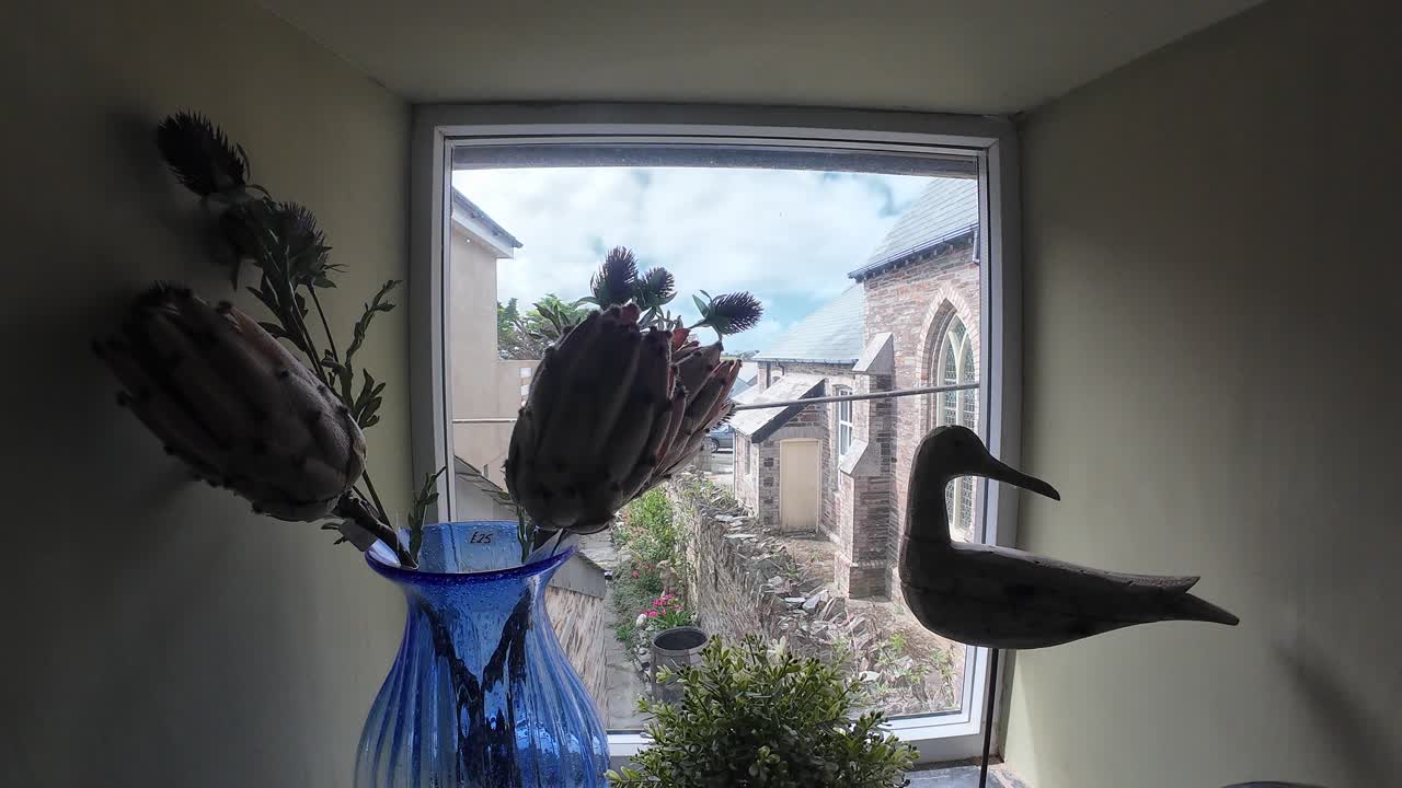 Delightful window view showcasing rustic decor, with a vibrant blue vase and wooden bird silhouette, framed by Cornwall architecture. Bright natural light fills this inviting scene