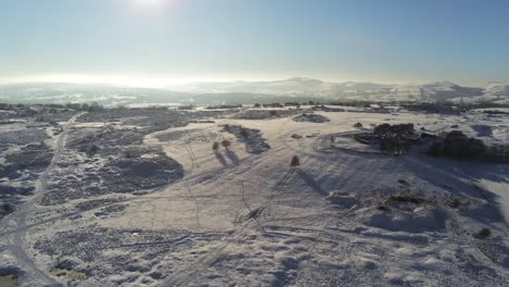 Snow-covered-rural-winter-countryside-track-footprint-shadows-terrain-aerial-view-lowering-left