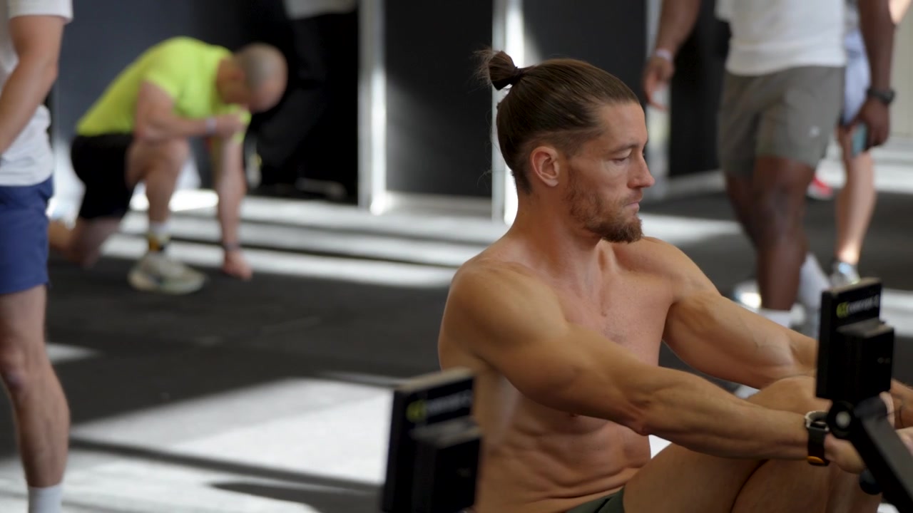 Gros plan intense d'un athlète musclé torse nu avec un chignon masculin tirant une poignée de RowErg avec détermination lors d'une séance d'échauffement Hyrox dans une salle de sport animée.