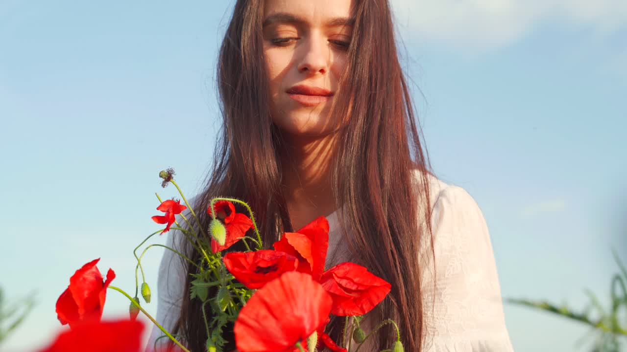 joven mujer caucásica con cabello castaño largo y ramo se inclina para oler la flor de amapola roja en el campo, de mano cerca