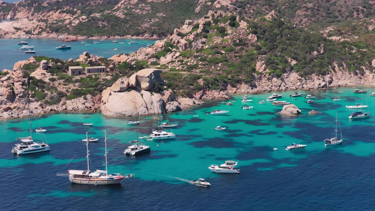 Vue aérienne au téléobjectif de yachts à voile de luxe et de bateaux de tourisme arrivant dans un port aux eaux turquoise niché parmi des falaises de granit sur l'île de Spargi dans l'archipel de la Maddalena en Sardaigne, Italie
