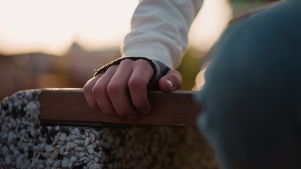 mano caucasica appoggiata su una panchina, guanto senza dita, ginocchio in denim, calda retroilluminazione del tramonto, profondità di campo ridotta, chitarrista da strada che si riscalda, crepuscolo nel parco urbano, bracciolo in cemento ruvido
