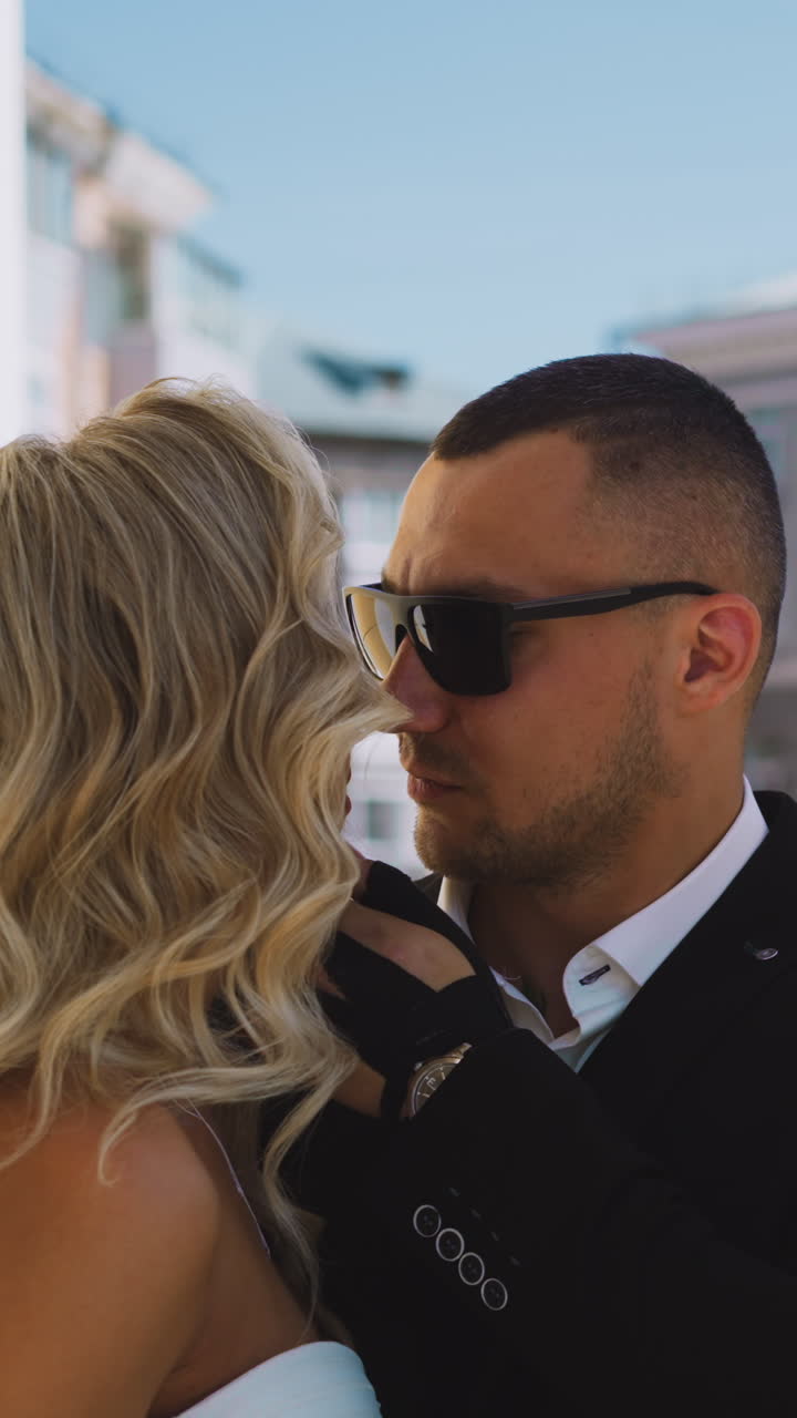 Confident man in suit with sunglasses touches cheek of beloved young bride with elegant hairdo standing by columns on street on summer day slow motion