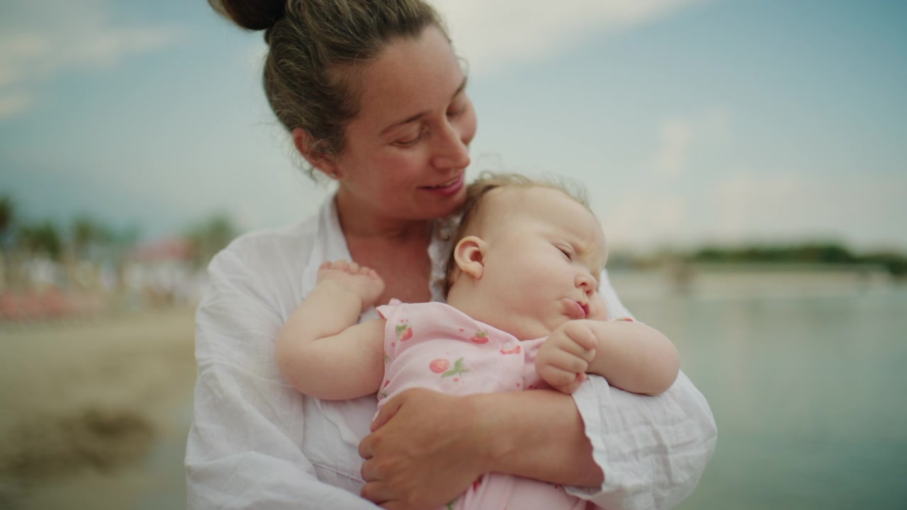 Neugieriges Baby lehnt sich vor, während es von einer Mutter nahe der Küstenlinie gehalten wird, und beobachtet sanfte Meereswellen während eines Familienstrandurlaubs.