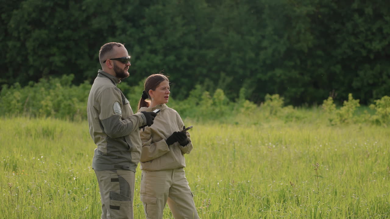 field worker uses handheld device while scanning distant aerial target in tall grassy field under bright sky near forest edge combining technology and outdoor research for environmental