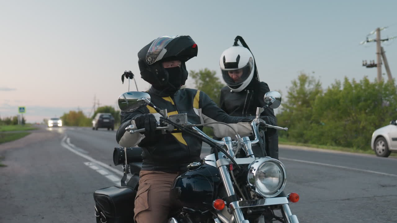 Two motorbike riders in helmets stop along roadside, one adjusting gear while another sits on chrome motorbike, as black car passes in blurred background
