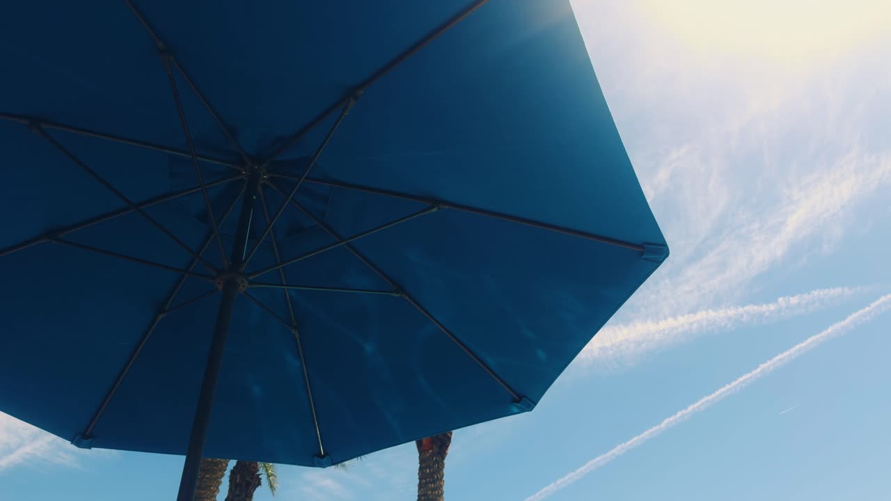 Looking up at a large blue beach umbrella under a bright sky with palm trees and light cloud streaks, on a warm and relaxing summer day