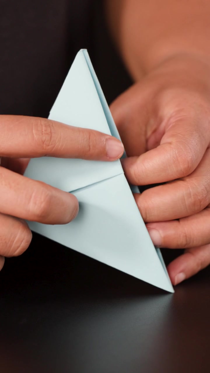 Primo piano delle mani che piegano con cura carta origami blu su una superficie scura con una morbida illuminazione da studio.