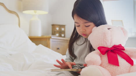 joven sentada en la cama con teléfono móvil con pijama en casa acurrucando juguete suave