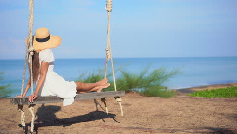 Mujer-Joven-En-Vestido-De-Verano-Y-Sombrero-Sentado-En-Un-Columpio-Con-Una-Vista-Impresionante-Del-Horizonte-Del-Mar-En-Un-Día-Soleado-De-Verano,-Fotograma-Completo-Estático