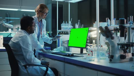 female research scientist has a conversation with bioengineer next to a desktop computer with green screen mock up. they look at computer template in a modern science laboratory.