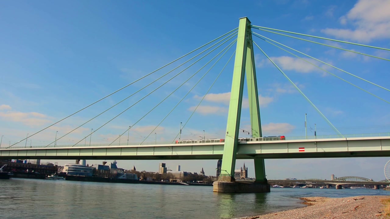 Wide river view in Cologne Germany featuring Severinsbrucke cable stayed bridge spanning Rhine River beneath bright blue sky with distant urban skyline