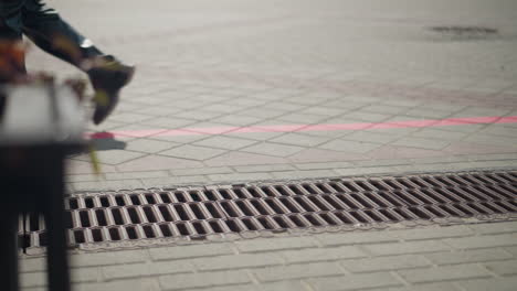 legs of passerby in black boot and trouser walking across interlocked pavement with a partially blurred view of flowers and a drainage grate in the foreground