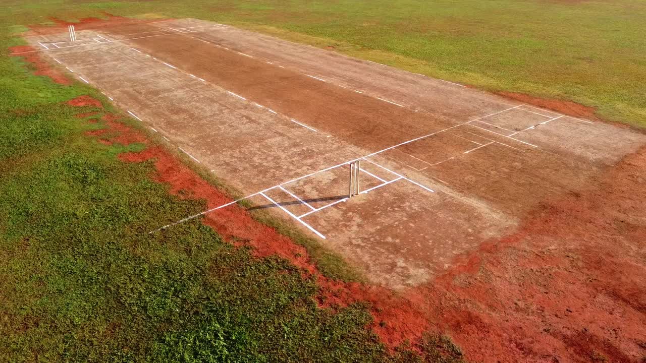 Ripresa aerea con drone di un campo da cricket rustico in terra battuta con linee bianche e paletti (wicket) in un campo verde rurale durante una giornata di sole