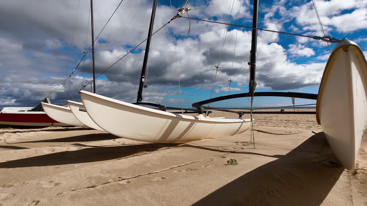 A white catamaran is anchored on a sandy beach, with its sails furled. The sky is a bright blue with fluffy white clouds. In Algarve, Portugal.