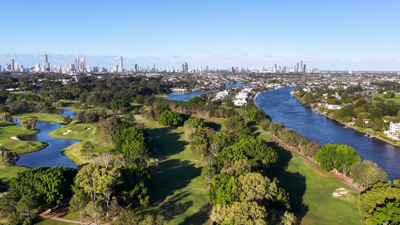 Ampia ripresa aerea con drone su un lussureggiante campo da golf e un fiume verso gli edifici dello skyline di una città distante.