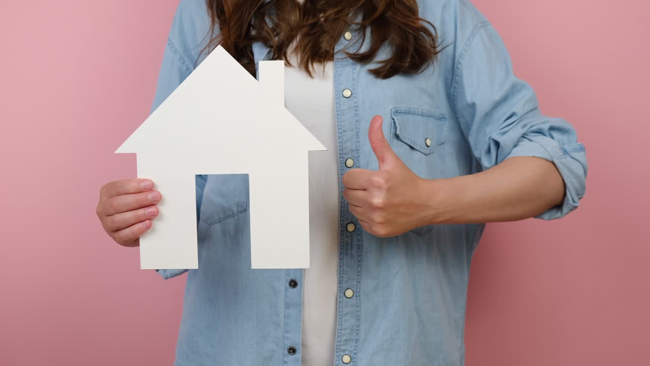 Close up of unrecognizable young woman pointing on white paper house model and makes like gesture, real estate home approval, isolated on pink studio background. Concept of social service about home